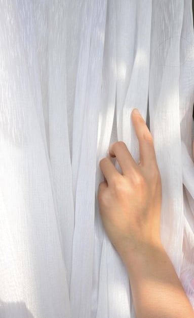 A close-up shot of a sheer white curtain gently swaying in the breeze. The sunlight filtering through the curtain creates a soft, diffused light, highlighting the texture of the fabric. The background is blurred, suggesting a window overlooking a garden or outdoor space.