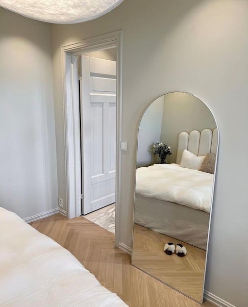 A modern bedroom with a full-length SHEIN mirror leaning against the wall. The mirror reflects the bed and other furniture, creating a sense of depth and spaciousness.